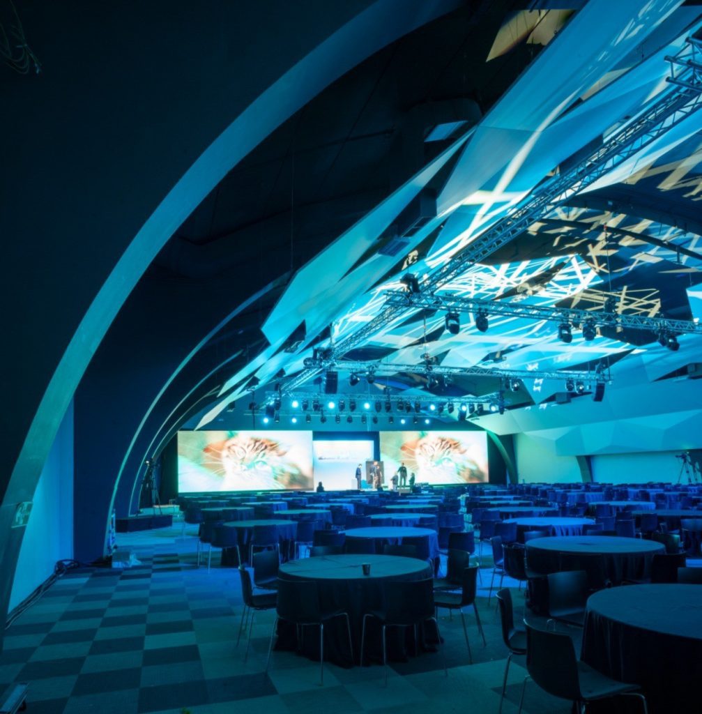 Sala de evento corporativo con pantallas LED panorámicas, iluminación azul y montaje técnico preparado antes del inicio.